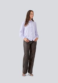 Helles lavendelfarbenes Hemd mit langen Ärmeln, kombiniert mit dunkelbraunen Lederhosen. Das Model steht in blauen Riemchensandalen vor einem grauen Hintergrund.