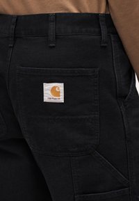 Črne kavbojke iz jeansa s velikim zadnjim žepom, na katerem je kvadratna bela etiketa z oranžnim logotipom. Tkanina ima gladko teksturo.