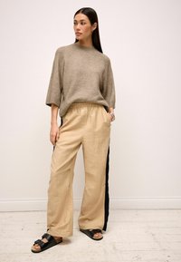 Lys beige striktrøje med korte ærmer og rund halsudskæring, parret med vide beige bukser med sorte sidelommer og sorte sandaler.