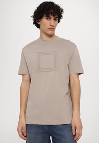 Beige bomulds t-shirt med rund hals, prydet med et hævet rektangulært logo design i midten, kombineret med blå denim jeans.