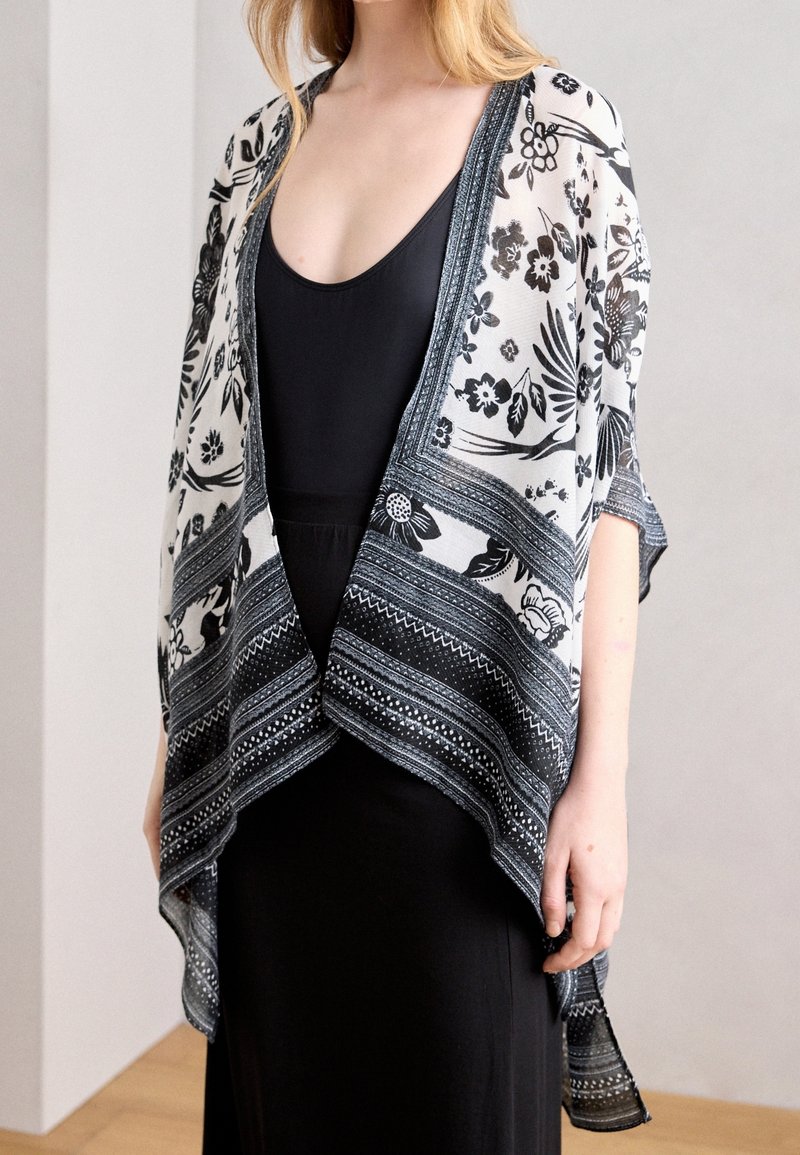 Femme portant une robe noire et un châle léger à motifs floraux noir et blanc drapé sur les épaules dans une pièce neutre.