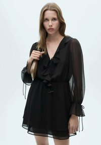 Black chiffon dress with ruffled neckline, long sleeves, and drawstring waist. Features tassel accents and a flowy, layered skirt.