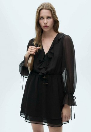 Black chiffon dress with ruffled neckline, long sleeves, and drawstring waist. Features tassel accents and a flowy, layered skirt.