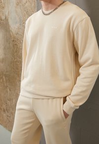 Sudadera beige con escote redondo y mangas largas, que presenta un detalle de logotipo minimalista. Combinada con pantalones de chándal beige a juego. Textura suave.