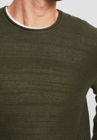 Pull en maille vert olive avec une finition texturée, col rond avec un col intérieur blanc, et fentes latérales pour faciliter le mouvement.