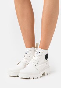 Bottes blanches en toile jusqu'à la cheville avec une semelle blanche épaisse et texturée, logo accent noir sur le côté, et fermeture à lacets sur le devant.