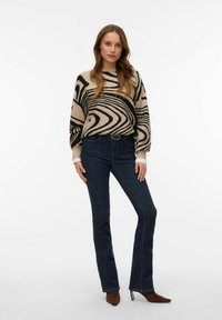 Béžový pulovr se černým vlnitým vzorem, doplněný tmavě modrými rozšířenými džíny. Outfit je doplněn páskem a hnědými špičatými podpatky.