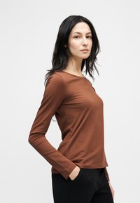 Γυναίκα με μακριά σκουρόχρωμα μαλλιά, ντυμένη με καφέ μακρυμάνικο πουκάμισο και μαύρο παντελόνι, στέκεται με το ένα χέρι στην τσέπη μπροστά σε λευκό φόντο.