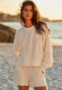 Lys beige bomulds sweatshirts med raglanærmer, parret med matchende shorts med let scallop kant. Blødt, tekstureret stof.