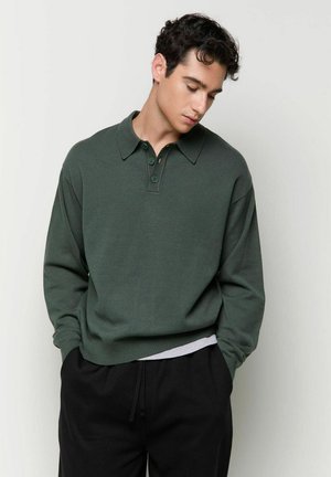 Junger Mann mit dunklem lockigen Haar, trägt ein dunkelgrünes Polo mit langen Ärmeln und schwarze Hose, schaut nach unten und hat die Hände in den Taschen.