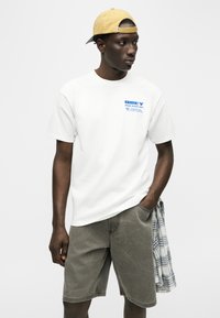 Νέος άνδρας που φοράει κίτρινο καπέλο με ανάποδη φορά, λευκό μπλουζάκι με γραφικά, γκρι σορτς και καρό πουκάμισο κρεμασμένο από την τσέπη, κοιτάζοντας προς τα δεξιά.