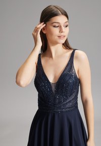 Abito da sera blu navy con un corpetto a V decorato con perline e dettagli trasparenti, accompagnato da una gonna fluida che mette in risalto intricate lavorazioni di perline e una texture liscia.