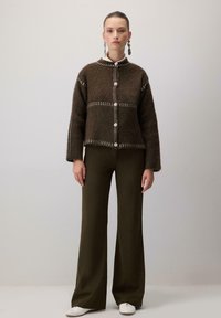 Femeie îmbrăcată cu un cardiguș fuzzy maro cu butoni și cusături albe, pantaloni evazați verzi închiși, pantofi albi și cercei lungi din argint, stând.