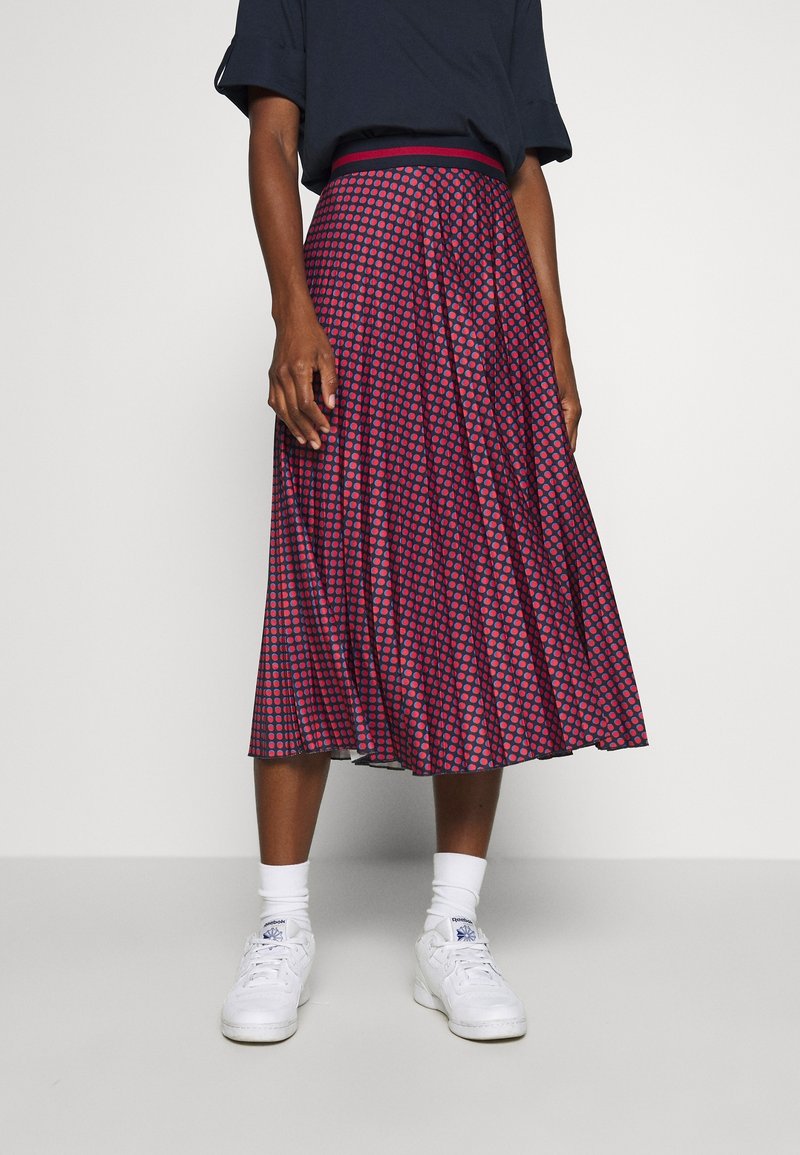Femme vêtue d'une jupe midi plissée à pois rouges et bleu marine, chaussettes blanches et baskets blanches, debout devant un fond uni.