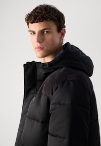 Jovem com cabelo curto e encaracolado, vestido com uma jaqueta acolchoada preta com capuz, a olhar para a câmara contra um fundo claro e liso.