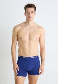 Barberløs mann med kort brunt hår iført blå Björn Borg boxershorts, stående mot en ensfarget hvit bakgrunn, vendt mot kameraet.
