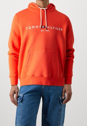 Personne portant un sweat à capuche Tommy Hilfiger orange vif avec des cordons blancs et un jean cargo bleu, debout devant un fond clair uni.