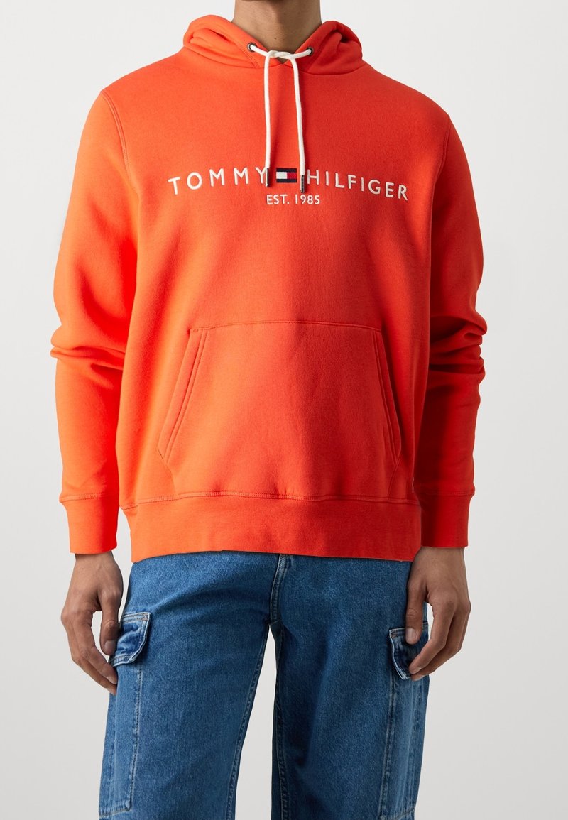 Personne portant un sweat à capuche Tommy Hilfiger orange vif avec des cordons blancs et un jean cargo bleu, debout devant un fond clair uni.