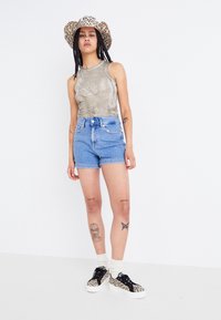 Top sin mangas de color verde oliva, pantalones cortos de mezclilla azul de cintura alta y zapatillas de leopardo en blanco y negro con suelas blancas. El sombrero tiene un patrón de leopardo.