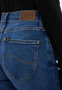 Temno modri denim kavbojki z visokim pasom, ki vključujejo usnjen znak z logotipom "Lee", opazno šivanje ter zadnje žepe s ukrivljenimi šivi.