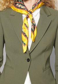 Blazer verde oliva con un design aderente, caratterizzato da una chiusura a un solo bottone. Accompagnato da una sciarpa di seta gialla con motivi floreali e accenti rossi.