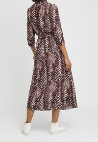 Maxiklänning i mönstrad tyg med röd, svart och vit zigzag-design; har en indragen midja och långa ärmar; matchas med vita sneakers.