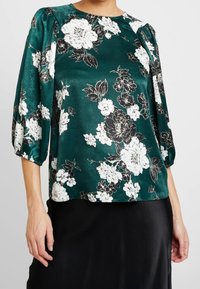 Teal floral blouse in a silky material with white and black blossoms, rounded neckline, and puffed elbow-length sleeves.