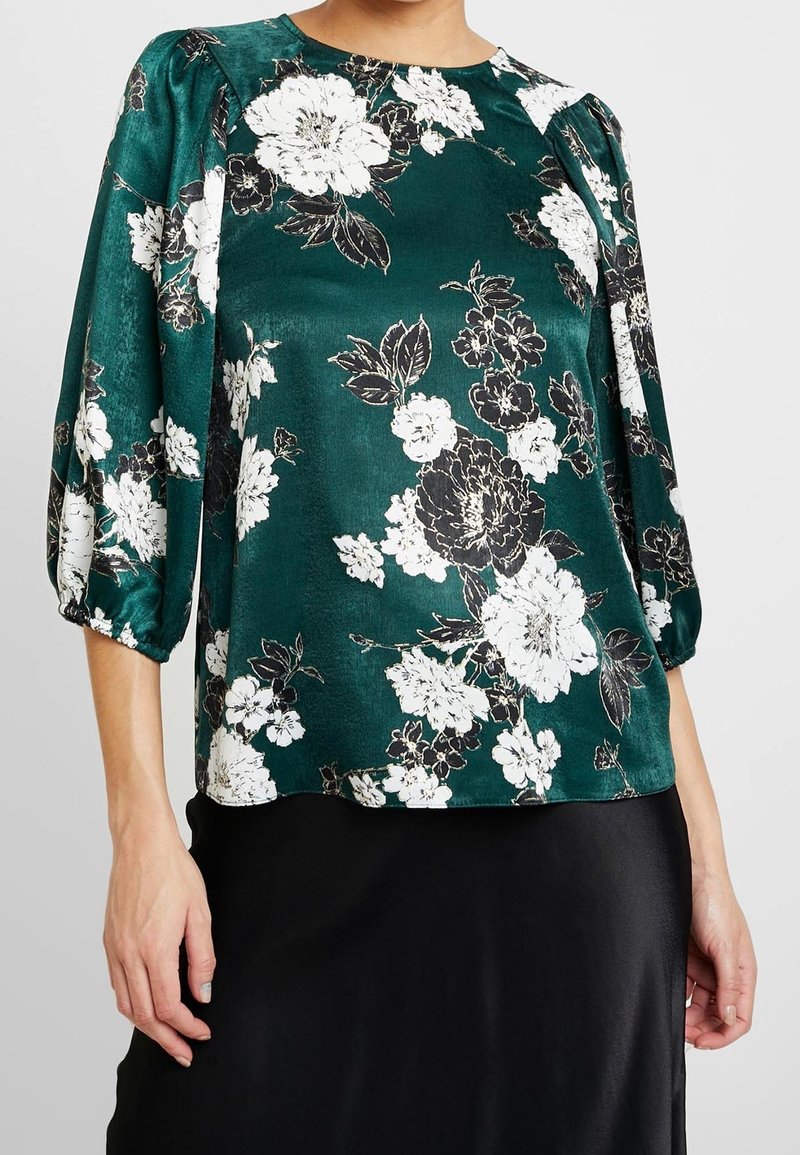 Teal floral blouse in a silky material with white and black blossoms, rounded neckline, and puffed elbow-length sleeves.
