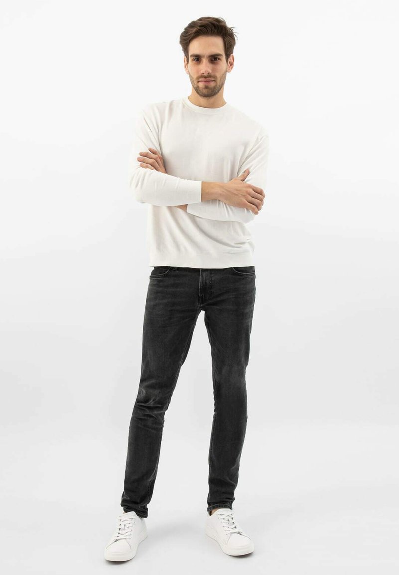 Weißes langärmliges Hemd, kombiniert mit schwarzen Slim-Fit-Jeans und weißen Sneakern. Das Outfit zeichnet sich durch eine glatte Textur und ein lässiges Design aus.