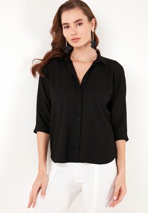Black button-up shirt with a collar and rolled sleeves, featuring a textured fabric. Worn with white pants. No visible patterns or accents.