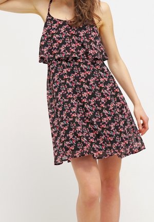 Kobieta w czarnej, bezrękawnikowej sukience do kolan z różowym kwiatowym wzorem i falbaną, stojąca na jednolitym białym tle.