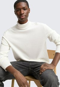 Weißer Strickkragenpullover mit gerippten Bündchen und Saum, kombiniert mit dunkelgrauen Jeans. Modell sitzt auf einem Holzstuhl vor neutralem Hintergrund.