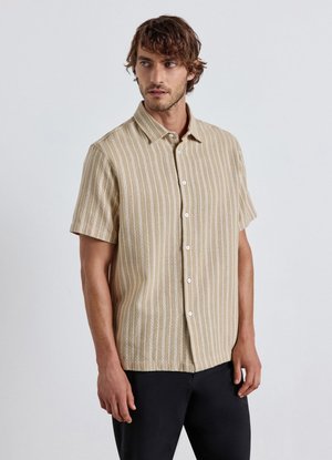 Mann trägt beige kurzärmeliges Hemd mit weißen vertikalen Streifen und schwarze Hose, steht vor einfarbigem Hintergrund.