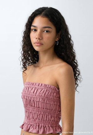 Jeune femme aux cheveux bouclés foncés et aux taches de rousseur, portant un top bustier rose froncé et des boucles d'oreilles anneaux argentées, regardant droit devant avec une expression neutre.
