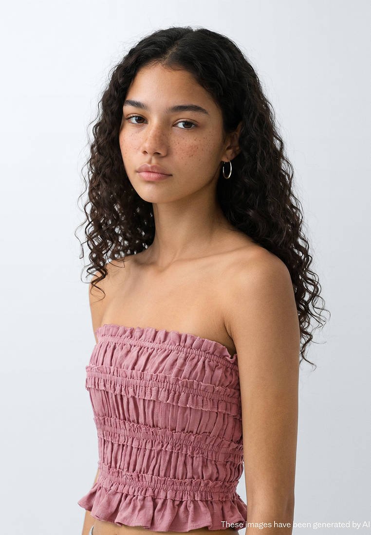 Jeune femme aux cheveux bouclés foncés et aux taches de rousseur, portant un top bustier rose froncé et des boucles d'oreilles anneaux argentées, regardant droit devant avec une expression neutre.