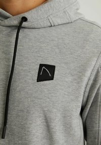 Acercamiento a una sudadera gris con cordones negros y un pequeño parche negro cosido con un diseño abstracto blanco en el pecho.