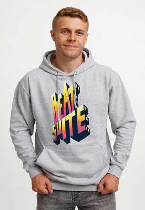Jeune homme souriant, portant un sweat à capuche gris avec un texte 3D coloré « BEAT SUITE » sur le devant, debout devant un fond blanc uni.