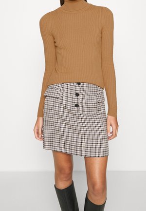 Femme portant un pull à col roulé côtelé beige, une jupe boutonnée devant motif pied-de-poule noir et blanc, et des bottes noires montant jusqu'aux genoux.