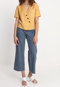 Camicia a maniche corte a righe gialle con scollo a V e taschino sul petto, abbinata a jeans a gamba larga blu con orli scuciti e sandali con tacco a blocco bianchi.