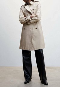 Beżowy trencz z podwójnym zapięciem, naramiennikami, wykonany z jasnego beżowego materiału, noszony na czarnych skórzanych spodniach i spiczastych butach.