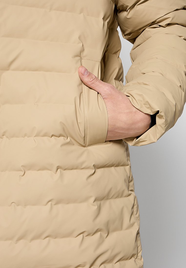 Doudoune beige avec un design matelassé, dotée d'une poche latérale et d'une texture douce et lisse. Le tissu semble léger et isolant.