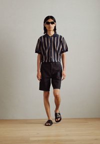 Kortermet marineblå skjorte med vertikale hvite striper, parret med svarte shorts og svarte sandaler. Modellen har på seg mørke solbriller.