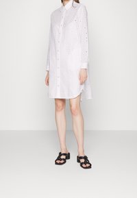 Vestido camisero blanco con botones, que presenta recortes de ojales, mangas largas y un dobladillo curvado. Incluye un bolsillo con parche en la parte delantera. Sandalias negras.