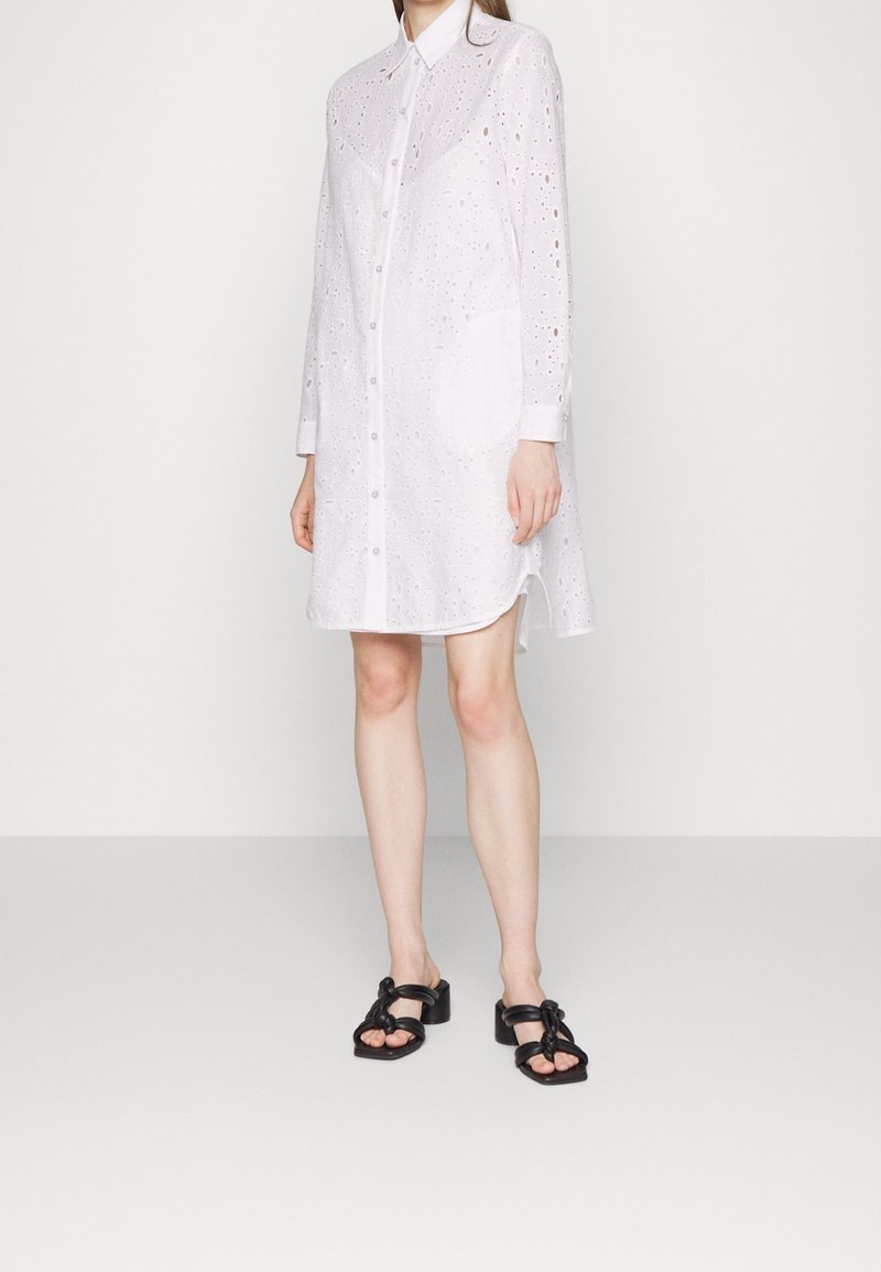 Vestido camisero blanco con botones, que presenta recortes de ojales, mangas largas y un dobladillo curvado. Incluye un bolsillo con parche en la parte delantera. Sandalias negras.