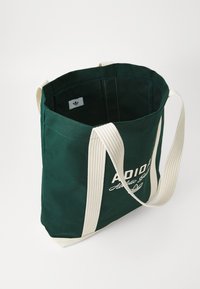 Dunkelgrüne Stofftasche mit weißen Griffen und Adidas-Logo in Weiß auf der Vorderseite, geöffnet gezeigt mit sichtbarem inneren Etikett.