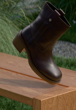 Donkerbruine leren enkellaars met een lage houten hak, rustend op een houten bank buiten, met gras en planten op de achtergrond.