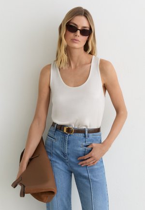 Mujer con cabello rubio hasta los hombros que lleva gafas de sol negras, top blanco sin mangas, vaqueros azul claro, sosteniendo una gran bolsa marrón.