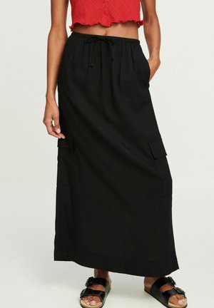Woman wearing a black long skirt with side pockets and a drawstring waist, a red cropped sleeveless top, and black buckle sandals.