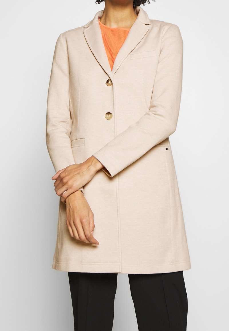 Cappotto beige su misura con rever a taglio, due bottoni dorati e tasche laterali. Presenta una texture liscia e una silhouette strutturata.