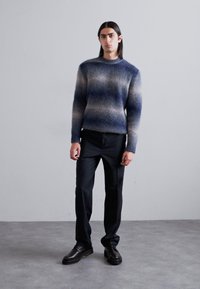 Pull en tricot dégradé de bleus et de gris, col rond, manches longues, associé à un pantalon sombre et des chaussures noires. Texture lisse, design ajusté.